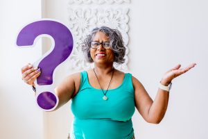 A brown skinned woman wearing a green vest top and standing in front of a white decorative wall. She is holding a purple question mark. 